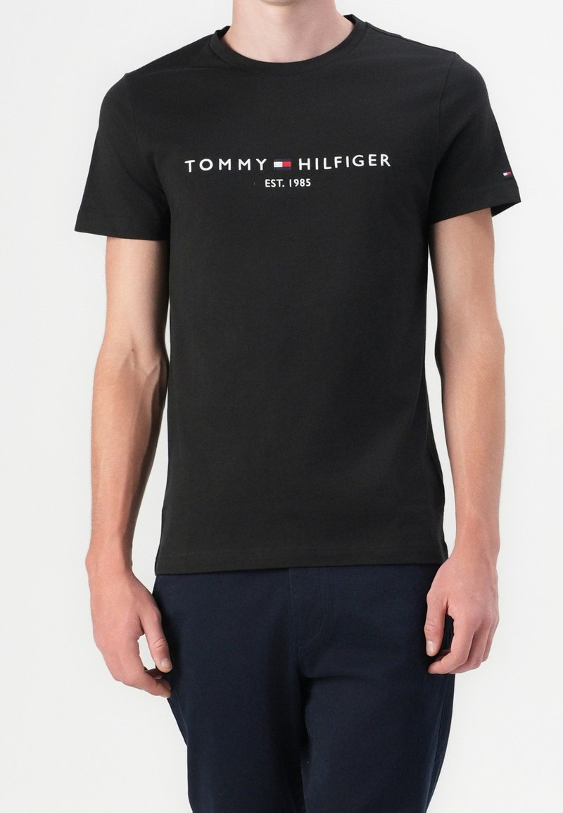 Černé bavlněné tričko s kulatým výstřihem, s logem "TOMMY HILFIGER EST. 1985" v bílé a červené barvě napříč hrudníkem. Krátké rukávy, minimalistický design.