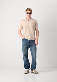 Knitted beige polo with short sleeves, textured pattern, paired with blue jeans and gray-brown sneakers. Accessories include dark sunglasses.