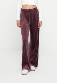 Frau trägt mauvefarbene Velour-Hose mit weitem Bein und Kordelzugbund sowie weiße Sneakers und ein kurzes, weißes Langarmoberteil.
