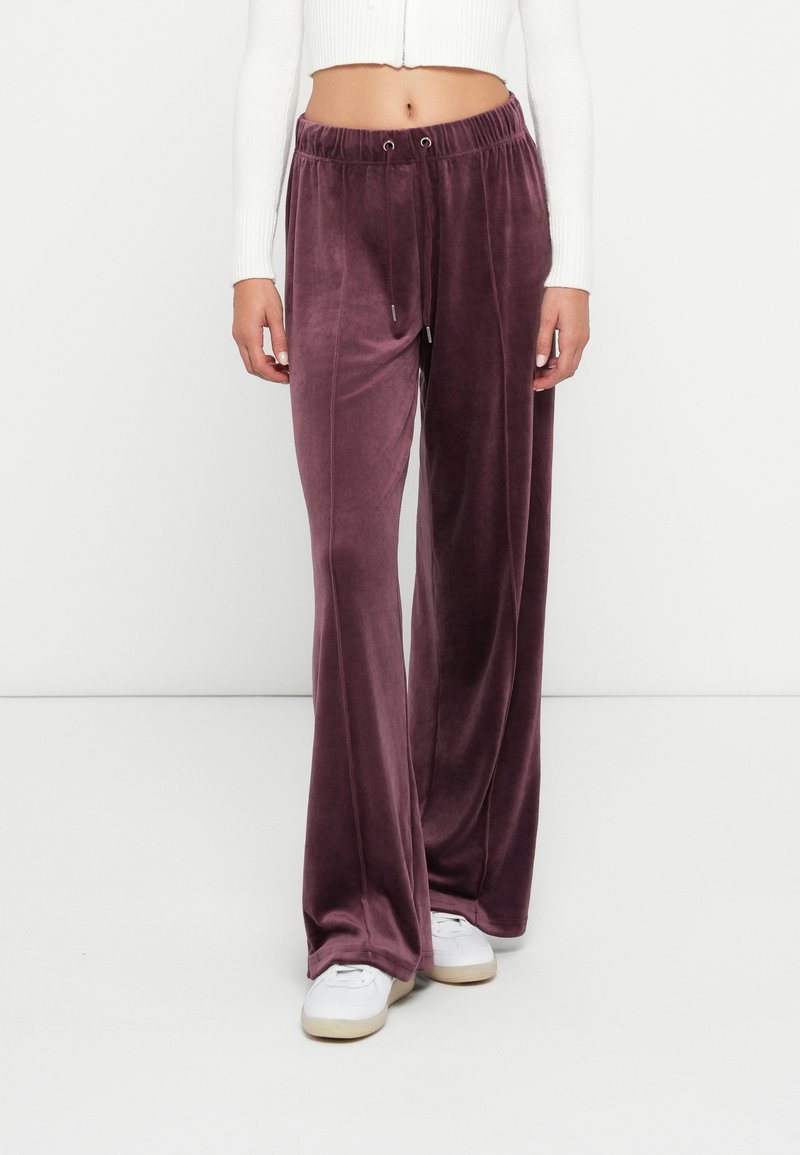 Frau trägt mauvefarbene Velour-Hose mit weitem Bein und Kordelzugbund sowie weiße Sneakers und ein kurzes, weißes Langarmoberteil.