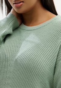 Maglione a maglia verde chiaro caratterizzato da una trama texture, scollatura rotonda e vestibilità rilassata. Dettagli di cucitura visibili lungo la parte frontale e le maniche.
