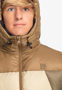 Bruine en beige gewatteerde jas met een hoge kraag, verstelbare trekkoorden en een logo patch. Gladde, gestikte textuur met duidelijke kleurvlakken.