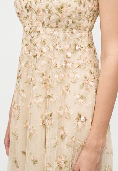 Μπεζ διάφανο φόρεμα με λεπτή ροζ και λευκή κεντημένη ανθοδέσμη και παγιέτες, φορεμένο από άτομο με εμφανή χέρι στα δεξιά.