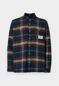 Camisa de flanela xadrez em azul marinho, castanho e creme. Apresenta fecho de botão, um bolso no peito e uma etiqueta com a marca na lateral.