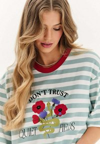 Žena s dlouhými vlnitými blond vlasy, která má na sobě zelené a bílé pruhované tričko s červeným límcem a květinovým hadím grafikem, na němž je nápis "NEDŮVEŘUJ TIŠE POLÍM".