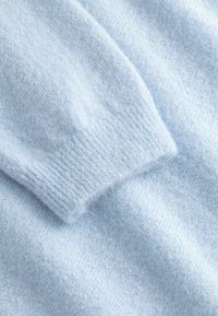 Hellblauer gestrickter Wollpullover mit Rippenbündchen am Ärmel, der auf einem passenden Stoffhintergrund liegt.