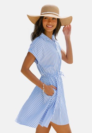 Donna sorridente, che indossa un vestito a maniche corte a righe blu e bianche con una cintura in vita, un cappello di paglia a tesa larga e un braccialetto di conchiglie.