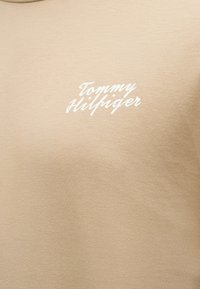 Μπεζ ύφασμα με λευκό καλλιγραφικό λογότυπο «Tommy Hilfiger» στο κέντρο της περιοχής του στήθους.