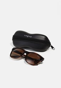Ochelari de soare cu tentă maro, cu rame groase, negre, așezați lângă un toc negru cu fermoar, etichetat "vogue". Design simplu, fără modele.