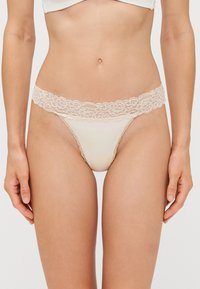 String en dentelle nude avec un corps en tissu doux, une taille en dentelle florale et un design à couverture minimaliste.