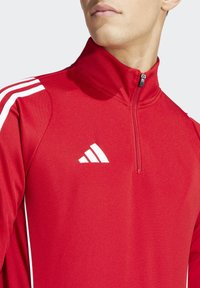 Red athletic zip-up jacket with white accents and three white stripes on the shoulders. Features a high collar and front zipper.
