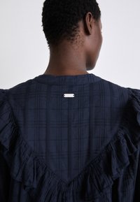Navyblaues Kleid mit V-förmigem Rücken, gerafften Akzenten und einem silbernen Logo-Detail. Der Stoff zeigt ein dezentes Karomuster.