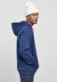Felpa con cappuccio blu in cotone. Presenta una tasca a marsupio e polsini a costine. Abbinata a un berretto di lana bianco. Vista laterale.