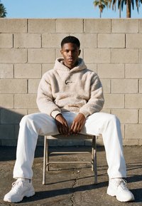 Ung mand iført hvid fleece-hoodie, hvide bukser og sneakers sidder med benene spredt på en metalstol mod en grå murstensvæg udendørs.