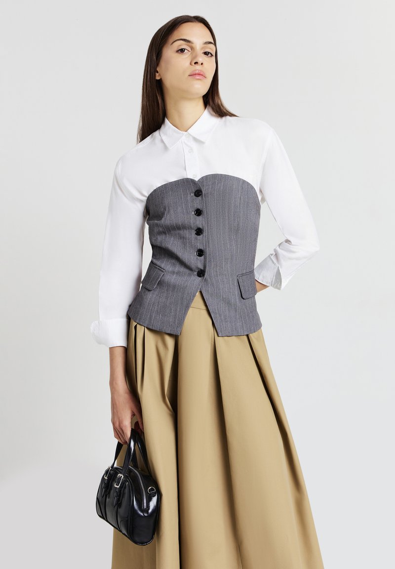 Donna che indossa una camicia bianca con un corsetto a righe grigie, una gonna plissettata beige, mentre tiene una piccola borsa nera su uno sfondo bianco.