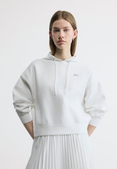Jeune femme aux cheveux lisses portant un sweat à capuche blanc et une jupe plissée blanche, debout les mains derrière le dos, sur fond uni.