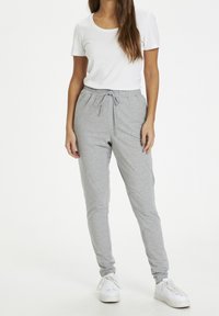 Vrouw in een witte t-shirt met korte mouwen, lichtgrijze joggers met trekkoord en witte sneakers, staand tegen een effen witte achtergrond.