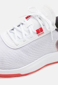 Sapatilha desportiva com um upper texturizado em cinza claro, atacadores brancos, detalhes em vermelho e uma sola branca com um vibrante solado vermelho.