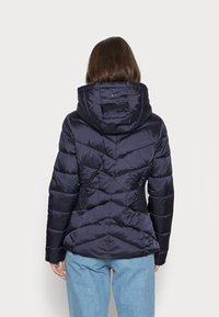 Doudoune bleu marine avec capuche, présentant un motif matelassé, des accents élastiques sur les côtés et une silhouette cintrée. Texture de tissu lisse et brillante.