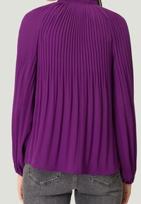 Violetti laskostettu paita, jossa on pitkät hihat ja korkea kaula-aukko. Valmistettu sileästä kankaasta, jossa on elastiset hihansuut ja lentävä siluetti.
