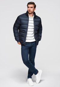 Marineblå pufferjakke med lynlås foran, parret med en hvid og marineblå stribet skjorte og mørkeblå jeans. Hvide sneakers fuldender looket.