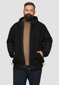 Schwarze Kapuzenjacke aus einem strukturierten Stoff mit Reißverschluss vorne. Zwei Seitentaschen und verstellbarer Kordelzug in der Kapuze, getragen über einem braunen Hemd.