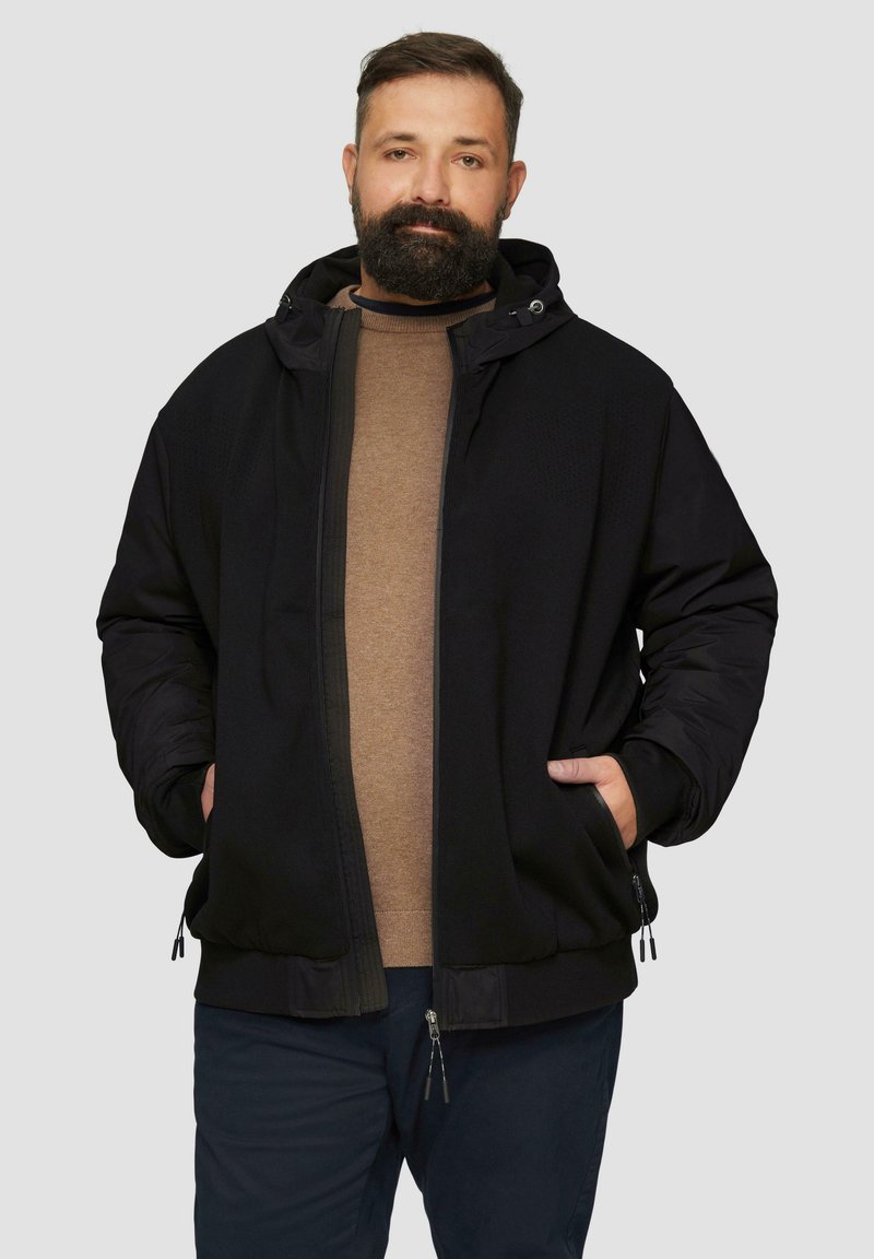 Schwarze Kapuzenjacke aus einem strukturierten Stoff mit Reißverschluss vorne. Zwei Seitentaschen und verstellbarer Kordelzug in der Kapuze, getragen über einem braunen Hemd.