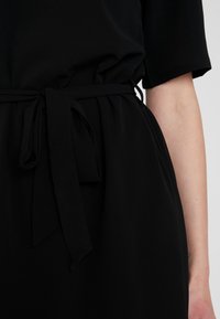 Zwarte jurk met een zachte textuur, een ontspannen pasvorm, korte mouwen en een strikdetail bij de taille. De stof lijkt lichtgewicht en vloeiend te zijn.