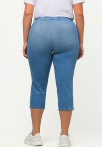 Jeans capri ajustados de cintura alta en denim azul claro con un ligero desgastado. Presentan trabillas para cinturón, bolsillos traseros y un dobladillo recortado. Se llevan zapatillas.
