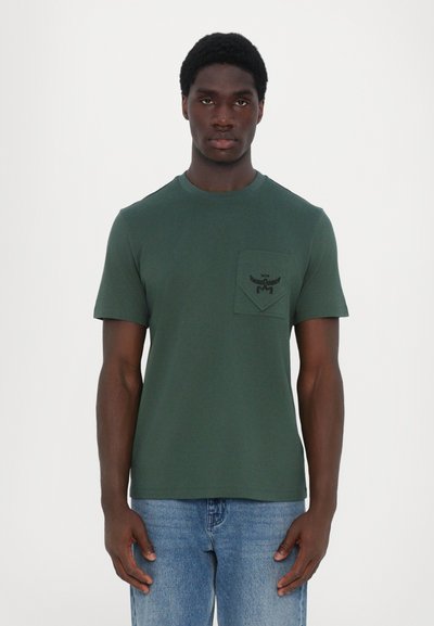 Camiseta de algodón verde con cuello redondo, mangas cortas y un bolsillo tipo parche. Presenta un detalle del logo en negro en el bolsillo.