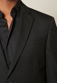 Veste en tissu noir texturé avec un col à revers cranté, une poche poitrine unique, et une chemise noire boutonnée en dessous.