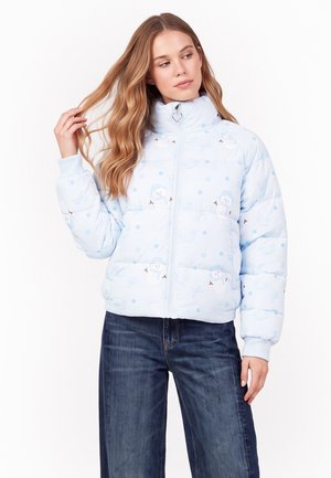 Vrouw die een lichtblauw donsjack met een sneeuwpopmotief draagt, staat met één hand haar aanrakend en kijkt opzij.