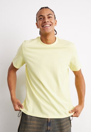 Camiseta amarilla de manga corta, hecha de algodón suave, con un cuello redondo y un pequeño logo en el pecho, combinada con pantalones oscuros a rayas.