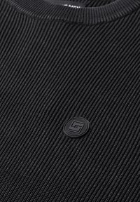 Tissu côtelé noir avec un petit emblème rond noir présentant un motif géométrique près du centre.