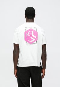 Vit t-shirt med korta ärmar, prydd med en stor rosa grafisk ikon och texten "KARL LAGERFELD JEANS" på baksidan. Materialet ser mjukt ut och passar för en avslappnad stil.