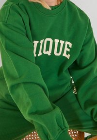 Sudadera verde con puños acanalados, que presenta la palabra "UNIQUE" en letras crema en el pecho. Tejido suave y texturizado.