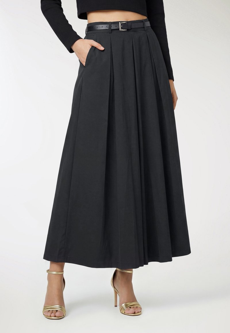 Kobieta ubrana w czarną plisowaną maxi spódnicę z wysokim stanem, z czarnym paskiem i złotymi sandałami na wysokim obcasie, stojąca z jedną ręką w kieszeni.