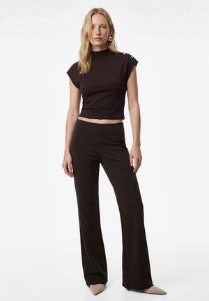 Vrouw met lang blond haar die een zwarte mouwloze mockneck top met knopen draagt en zwarte wijde broek met beige puntige schoenen.