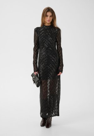 Jeune femme portant une longue robe noire à sequins et tenant une pochette noire, debout devant un fond clair uni.