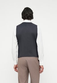 Colete azul-marinho com uma textura suave, usado sobre uma camisa branca. As costas são lisas, combinado com calças castanhas claras que apresentam um corte ajustado.