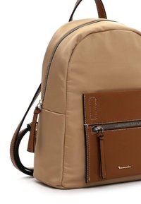 Sac à dos en tissu beige avec un haut incurvé, poche avant en cuir marron, fermeture éclaire argentée et détail de logo. Bretelles réglables et design robuste.