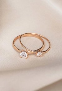 Zwei Ringe aus Roségold mit klaren runden Edelsteinen, die eine glatte Oberfläche und ein minimalistisches Design aufweisen, liegen auf einer hellen Stoffoberfläche.