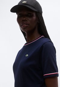 Tumši zila īsroku krekls ar baltu un sarkanu svītru apkakli, kreisajā krūtīs zaļš krokodila logotips. Kombinēts ar melnu cepuri.