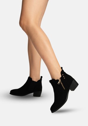 Benen met zwarte suède enkellaarsjes met lage blokhakken en gouden ritsdetail, geplaatst tegen een effen lichte achtergrond.