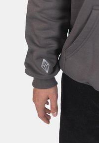 Felpa grigia con polsini a coste; presenta un logo geometrico bianco ricamato sulla manica sinistra; tessuto morbido e testurizzato.