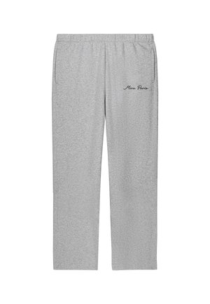 Jonge vrouw staat met handen in de zakken, draagt een oversized wit T-shirt, grijze joggingbroek met de tekst "Mina Paris" en witte sneakers.