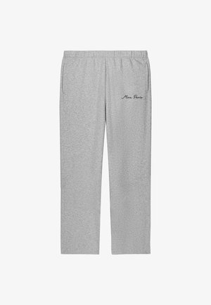 Jeune femme debout, les mains dans les poches, portant un t-shirt blanc oversize, un pantalon de survêtement gris avec le texte « Mina Paris » et des baskets blanches.