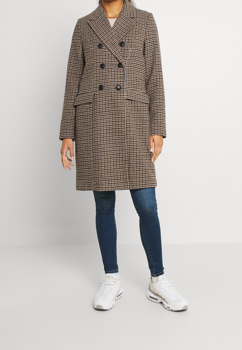 Personne portant un manteau croisé à carreaux marron et noir, un jean skinny bleu foncé et des baskets blanches, debout devant un fond uni.