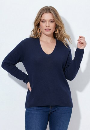 Kvinde iført en mørkeblå V-hals sweater og blå jeans, poserende med den ene hånd på hoften og den anden tæt ved hovedet.