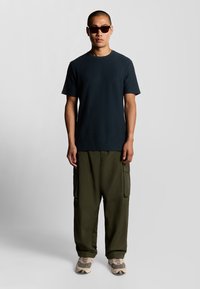 Zwart T-shirt met korte mouwen, losse olijfgroene cargobroeken met zijzakken en beige sneakers. Draag met donkere zonnebril.